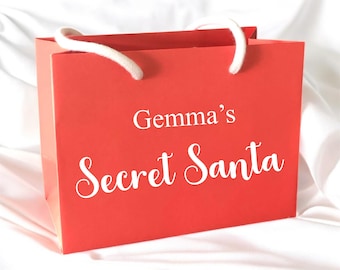 May include: A red gift bag with white rope handles. The text "Gemma's Secret Santa" is printed in white script on the front of the bag. The bag is sitting on a white, textured surface.