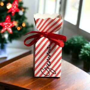 May include: A red and white striped candy cane shaped gift box with a red ribbon and the name "Gemma" written on the front.
