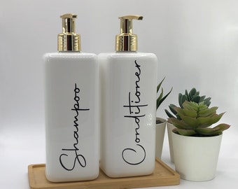 May include: Two rectangular white plastic bottles with gold-colored pump dispensers. The bottles are labeled with black cursive text reading Shampoo and Conditioner. They sit on a rectangular bamboo tray next to a small white pot with a succulent.