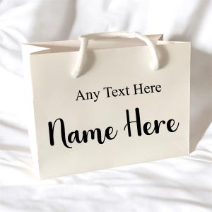 May include: A cream-coloured gift bag with white rope handles. The bag features the text "Any Text Here" in a simple font and "Name Here" in a cursive font, both in black. The bag is sitting on a white surface.