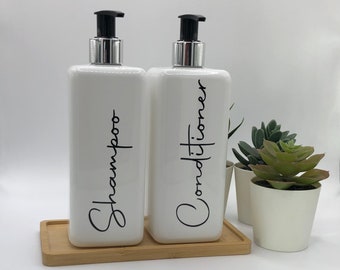 May include: Two rectangular white pump bottles with black script text reading Shampoo and Conditioner. The bottles feature silver collars and black pump heads, resting on a rectangular bamboo tray next to a small white pot with a green succulent.