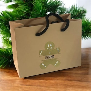 May include: A brown paper gift bag with black rope handles. The bag features a gold gingerbread man design with the name "Gemma" printed below. The background includes green pine branches.