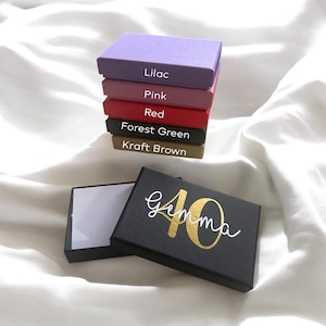 Pode incluir: Uma pilha de caixas de presente coloridas em lilás, rosa, vermelho, verde floresta e castanho kraft. Uma caixa preta aberta com o nome "Gemma" e o número "40" em dourado.