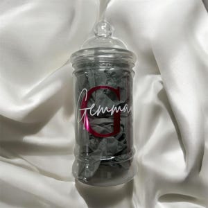May include: A clear glass apothecary jar with a lid, filled with grey tissue paper. The jar has a pink letter "G" and the name "Gemma" in white script. The jar sits on a white satin surface.