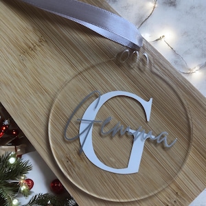 Könnte beinhalten: Ein runder, transparenter Acryl-Anhänger mit einem weißen Buchstaben G und dem Namen "Gemma" in silberner Schrift.