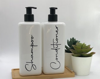 May include: Two rectangular white plastic bottles with black pump dispensers, each labeled with black cursive text reading Shampoo and Conditioner. The bottles sit on a rectangular bamboo tray next to a small white pot containing a green succulent.