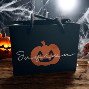 May include: Black gift bag with a jack-o'-lantern design and the name "Jayson" in white script. The bag has black rope handles and is set against a Halloween-themed background with spiderwebs and pumpkins.