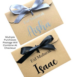 May include: Two kraft paper gift envelopes, one with a silver ribbon and the name "Aisha", the other with a black ribbon and the name "Isaac". Both envelopes have "Eid Mubarak" printed on them.