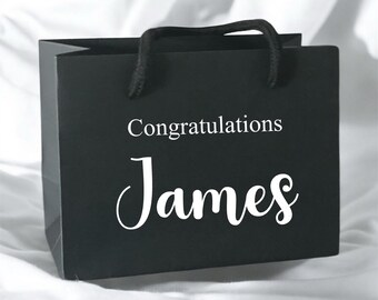 May include: A black gift bag with black rope handles. The bag has the words "Congratulations James" printed in white script. The bag is sitting on a white surface.