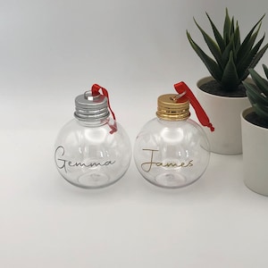 May include: Two clear plastic Christmas baubles with red ribbon hangers. The bauble on the left has a silver lid and the name "Gemma" written in gold lettering. The bauble on the right has a gold lid and the name "James" written in gold lettering.