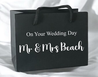 May include: A black gift bag with black rope handles featuring the white printed text On Your Wedding Day and Mr & Mrs Beach. The bag has a rectangular shape and a matte finish, displaying clear white lettering centered on the front panel.
