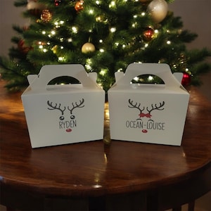 May include: Two white gift boxes with handles, each decorated with a reindeer face and a name. The box on the left says "RYDEN" and the box on the right says "OCEAN-LOUISE". The boxes are on a dark wooden surface, with a Christmas tree in the background.