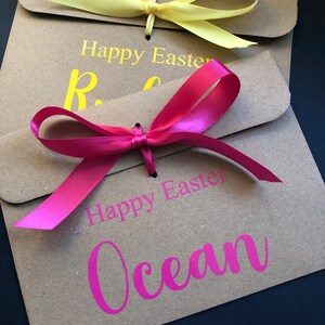 K&ouml;nnte beinhalten: Zwei braune Papiert&uuml;ten mit gelben und rosa Schleifen. Auf den T&uuml;ten stehen die Worte "Frohe Ostern" und Namen in Gelb und Pink. Der Hintergrund ist dunkel.
