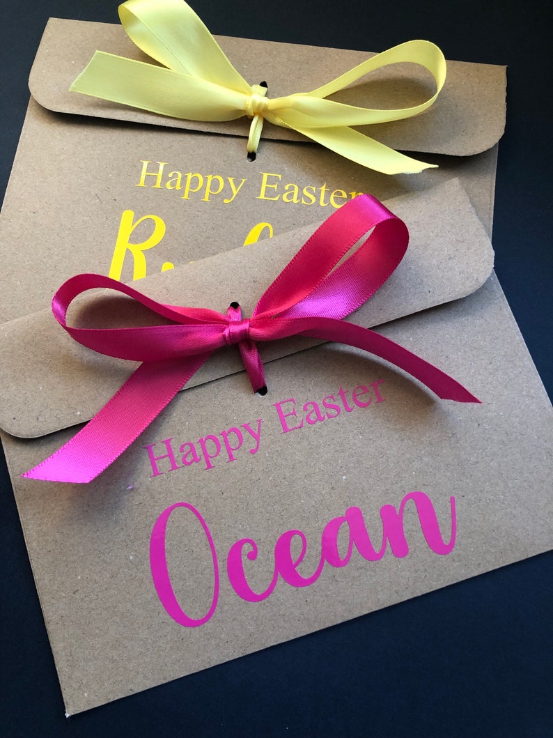 K&ouml;nnte beinhalten: Zwei braune Papiert&uuml;ten mit gelben und rosa Schleifen. Auf den T&uuml;ten stehen die Worte "Frohe Ostern" und Namen in Gelb und Pink. Der Hintergrund ist dunkel.