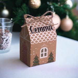Könnte beinhalten: Braune Geschenkbox aus Pappe in Form eines Lebkuchenhauses, mit dem Namen "Gemma" auf dem Dach. Die Box hat ein weißes, wellenförmiges Dachdesign, ein weißes Fenster und zwei grüne Weihnachtsbäume. Eine Kordelschleife ziert die Oberseite.