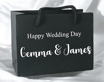 May include: A black gift bag with black rope handles featuring the white printed text Happy Wedding Day and the names Gemma & James in a script font. The bag has a rectangular shape and is constructed from a matte material with a clean finish.