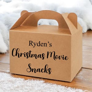 Può includere: Una scatola di cartone marrone con manico, con l'etichetta "Ryden's Christmas Movie Snacks" in corsivo nero. La scatola è su una superficie di legno con una spolverata di polvere bianca, forse neve. La scatola è progettata per trasportare snack.