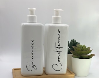 May include: Two rectangular white plastic pump bottles labeled Shampoo and Conditioner in black cursive script. The bottles sit on a rectangular light wood tray next to a small white pot containing a green succulent plant on a white background.