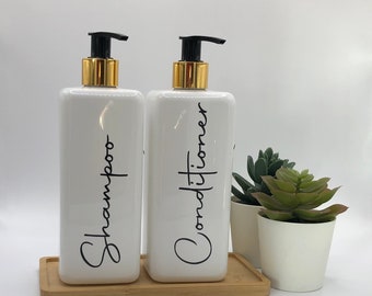 May include: Two rectangular white plastic bottles with black pump dispensers and gold collars. The bottles are labeled with black cursive text reading Shampoo and Conditioner. They sit on a rectangular bamboo tray next to two small potted succulents.