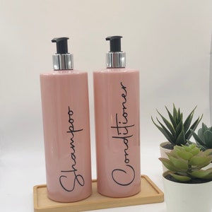 May include: Two pink shampoo and conditioner bottles with black text and silver pump tops on a wooden tray. The bottles are labelled "Shampoo" and "Conditioner". Small potted plants are in the background.