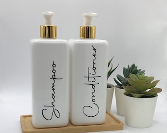 May include: Two rectangular white plastic bottles with gold pump dispensers, labeled Shampoo and Conditioner in black script. The bottles sit on a rectangular bamboo tray next to two small white pots containing green artificial succulent plants.