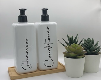 May include: Two white rectangular plastic bottles with black pump dispensers, labeled Shampoo and Conditioner in black script. The bottles sit on a rectangular bamboo tray next to two small green succulent plants in white pots.