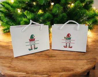 May include: Two white gift bags with white rope handles, each featuring a graphic of an elf hat and boots in red and green. The left bag displays the name RYDEN, and the right bag displays the name OCEAN, both printed in black text.