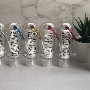 Pode incluir: Cinco garrafas de viagem de plástico transparente com mosquetões. As garrafas são etiquetadas com "Shampoo", "Condicionador", "Gel de banho", "Lavagem facial" e "Creme facial".