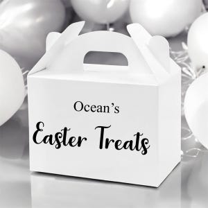 Puede incluir: Una caja blanca con asa, con el texto "Ocean's Easter Treats" en escritura negra. La caja está rodeada de globos blancos, creando un ambiente festivo y celebratorio.