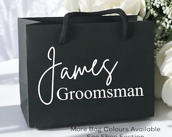 May include: A matte black gift bag with black rope handles. The front features the white text James in a cursive script font above the word Groomsman in a clean sans-serif font. The bag is positioned against a backdrop of white flowers.
