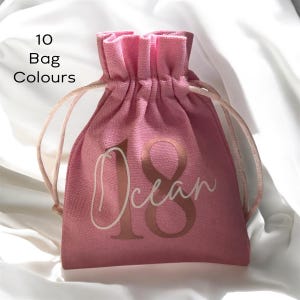 May include: A pink drawstring bag with the word "Ocean" and the number "18" printed on the front in a metallic gold color. The bag is made of a textured fabric and has a drawstring closure. The text "10 Bag Colours" is printed in black.