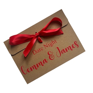 May include: A brown paper envelope tied with a red satin ribbon. The words "Date Night" and "Gemma & James" are printed in red script on the front. The envelope is on a white background.