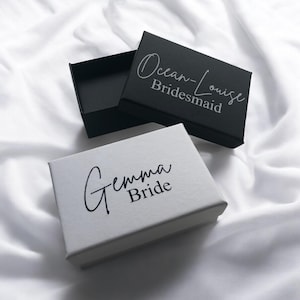 May include: Two small gift boxes, one black and one white, are displayed on a white, textured surface. The black box lid is open. The white box has the words "Gemma Bride" printed on it. The black box has the words "Ocean-Louise Bridesmaid" printed on it.