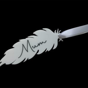 May include: A white feather-shaped ornament with the word "Mum" in black script. A white satin ribbon is attached to the top. The ornament is against a black background.