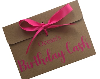 May include: A brown paper envelope tied with a bright pink ribbon. The words "Ocean's Birthday Cash" are printed in pink script on the front of the envelope. The envelope is closed with a pink ribbon tied in a bow.