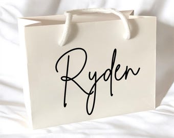 May include: Ivory gift bag with white rope handles. The name "Ryden" is written in a black cursive font on the front of the bag. The bag is sitting on a white surface.