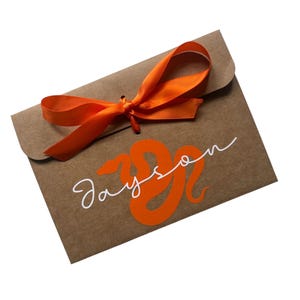 May include: A brown kraft paper envelope featuring a bright orange satin ribbon tied in a bow at the top flap. The front of the envelope displays a large orange graphic of a snake coiled into the shape of the letter S, with the name Jayson written in white cursive script across the centre.