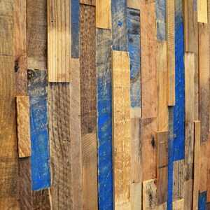 Op de afbeelding: Een muur gemaakt van gerecycled hout, geschilderd in verschillende tinten bruin en blauw. De planken zijn in een willekeurig patroon gerangschikt, waardoor een rustieke en industri&euml;le uitstraling ontstaat.