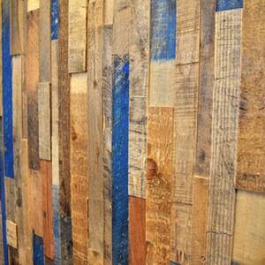 Op de afbeelding: Een muur bedekt met gerecyclede houten planken, waarvan sommige blauw geverfd zijn. De houten planken zijn in een willekeurig patroon gerangschikt, wat een rustieke en industri&euml;le uitstraling cre&euml;ert.