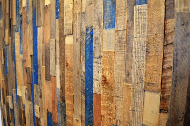 Op de afbeelding: Een muur bedekt met gerecyclede houten planken, waarvan sommige blauw geverfd zijn. De houten planken zijn in een willekeurig patroon gerangschikt, wat een rustieke en industri&euml;le uitstraling cre&euml;ert.