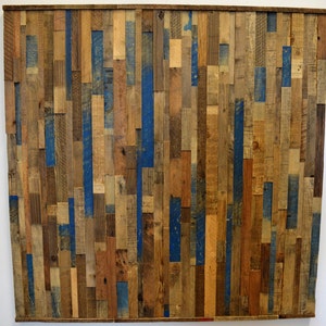 Op de afbeelding: Een wandkunstwerk gemaakt van gerecycled hout in verschillende tinten bruin en blauw. De planken zijn verticaal gerangschikt, waardoor een textuur en een rustieke uitstraling ontstaat.