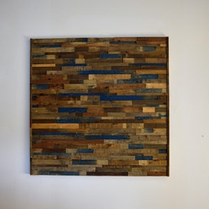 Op de afbeelding: Een wandkunstwerk gemaakt van gerecycled hout met een rustieke uitstraling. Het hout is in een horizontaal patroon gerangschikt en geschilderd in tinten bruin, blauw en beige.