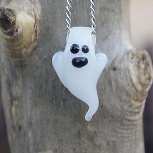 May include: A white glass ghost pendant with black eyes and a mouth hangs from a silver chain. The ghost is a whimsical and spooky Halloween decoration.