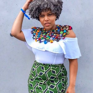 May include: A woman with gray hair is wearing a white off-the-shoulder top, a green and black patterned skirt, and a large colorful beaded necklace.