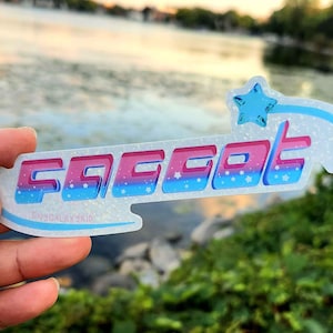 May include: A sparkly blue and pink sticker with the word "FGCOT" in a retro font. The sticker has a white background with a blue star and a blue comet tail.