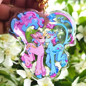 May include: A clear acrylic keychain featuring two cartoon ponies kissing. The ponies are pink and blue with colorful manes and tails. The keychain has a gold-colored clasp and a decorative design of pink and blue clouds and stars.