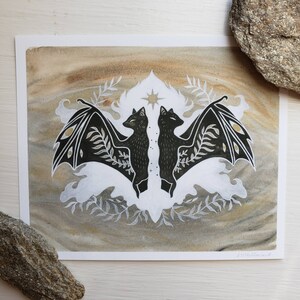 May include: A black and white illustration of two bats with wings spread, facing each other. The bats are surrounded by stylised foliage and a star in the centre. The background is a light brown watercolour wash.