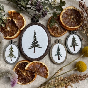 Könnte beinhalten: Eine Auswahl an gestickten Kunstwerken mit immergrünen Bäumen in ovalen Holzrahmen. Die Bäume sind grün und braun, vor einem weißen Hintergrund. Getrocknete Orangenscheiben und florale Elemente umgeben die gerahmte Kunst.