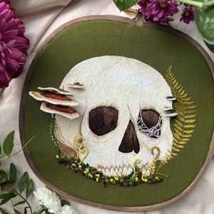 Op de afbeelding: Borduurwerk op een hoepel met een witte schedel met bruine ogen en een spinnenwebontwerp in één oog. De schedel is omringd door groen gebladerte, bruine paddenstoelen en gele bloemen.