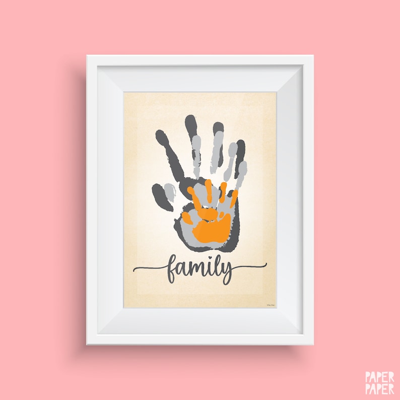 May include: A framed print featuring a handprint design in grey, light grey, and orange. The handprints are layered on top of each other, with the smallest handprint in the centre. The word "family" is written in cursive below the handprints.